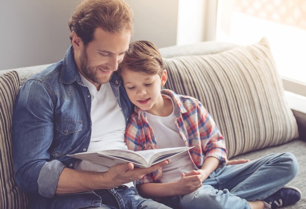 A man and his son sit together on a couch, enjoying a book during their parallel parenting time.