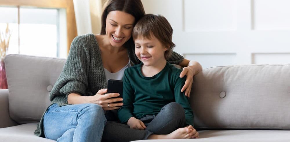 Loving young Caucasian mom and small son relax on couch in living room watch video on smartphone together.